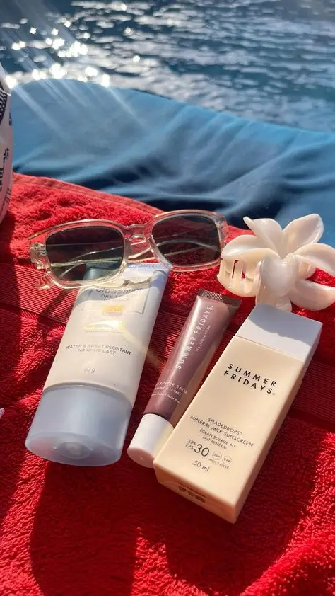 Poolside essentials: sunglasses, sunscreen, lip balm, hair clip on a red towel by the water. Ideal for a sunny summer day.