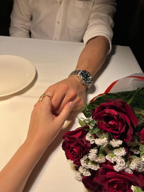 Couple holding hands over a romantic dinner table, with red roses and a watch visible.