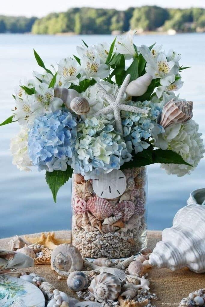 Seashell floral arrangement with hydrangeas, starfish, and shells in a vase by the water, perfect for beach-themed decor.