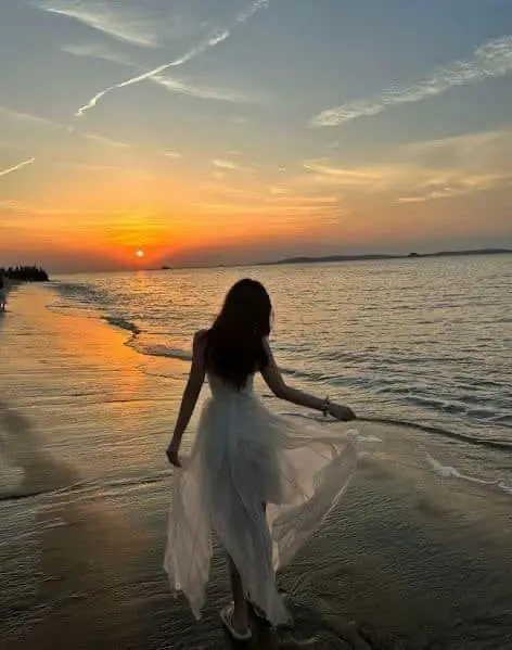 Woman in white dress walking on beach at sunset, with waves lapping at her feet. Vibrant sky with orange hues. Serene coastal scene.
