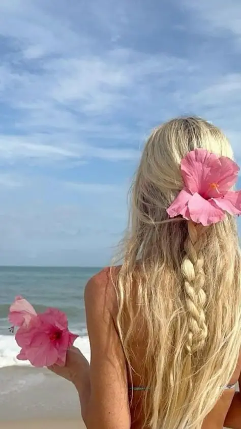 Blonde woman with braided hair and pink flowers at a beach, enjoying the ocean view under a blue sky.