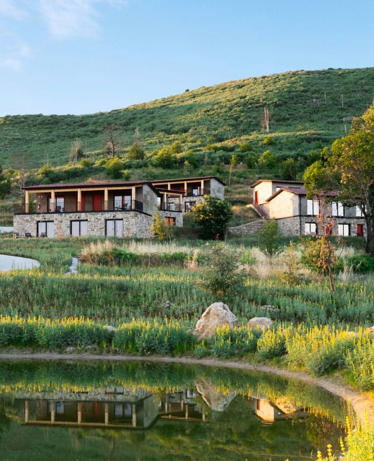 Stone villas on a lush hillside are reflected in a serene pond, under a clear blue sky. Ideal for a tranquil countryside escape.