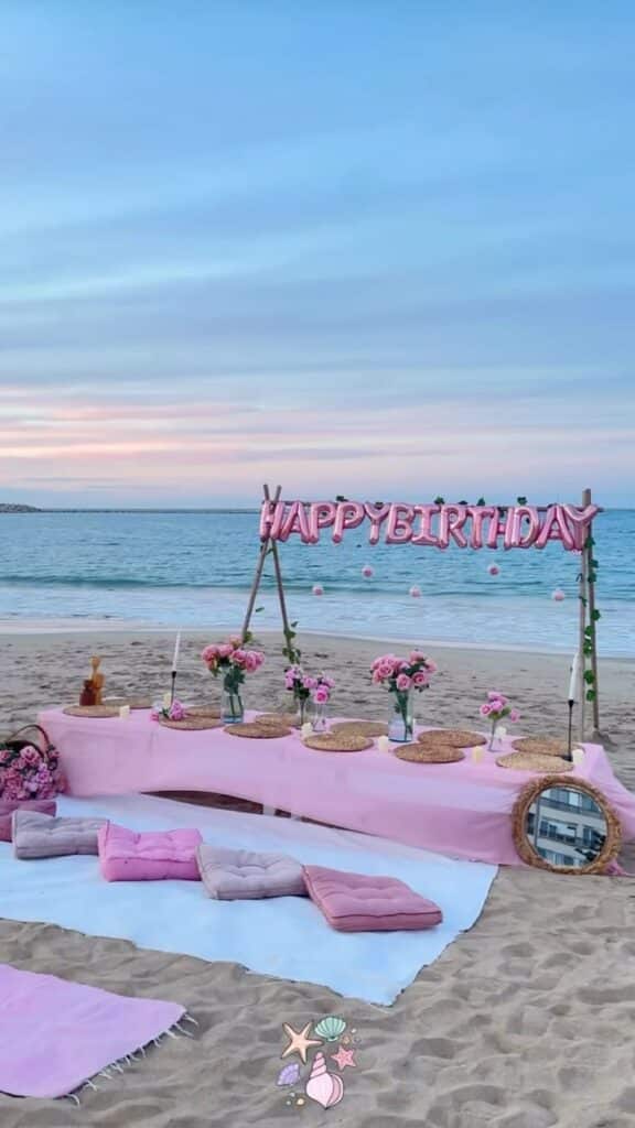 308d2ba0-455d-47c5-836a-1fcbe5f74969 - Handy Home Beach birthday picnic setup with pink decorations, flowers, cushions, and Happy Birthday balloons at sunrise.