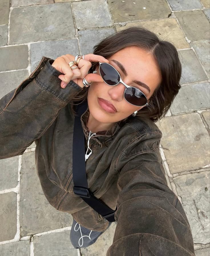 3f4bbdd7-0349-4cbc-b1ed-ac1289eed882 - Handy Home Woman in sunglasses taking a selfie outdoors, wearing a stylish leather jacket and accessories, on a stone pavement background.