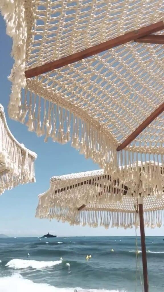 Crochet beach umbrellas with fringe shade a sunny seaside view, blue ocean waves, and a distant boat under a clear sky.
