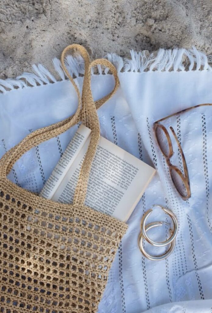 Woven beach bag with a book on a white blanket, sunglasses, and hoop earrings on sand, perfect for a summer day.