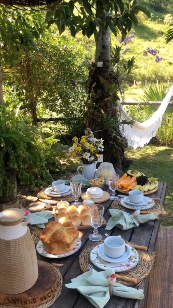 Outdoor brunch table set with pastries, fruits, and cups; lush garden and hammock in the background. Relaxing nature breakfast scene.