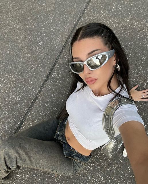 Woman in sunglasses and a white crop top taking a selfie from above, wearing jeans and a metallic backpack, on a textured surface.