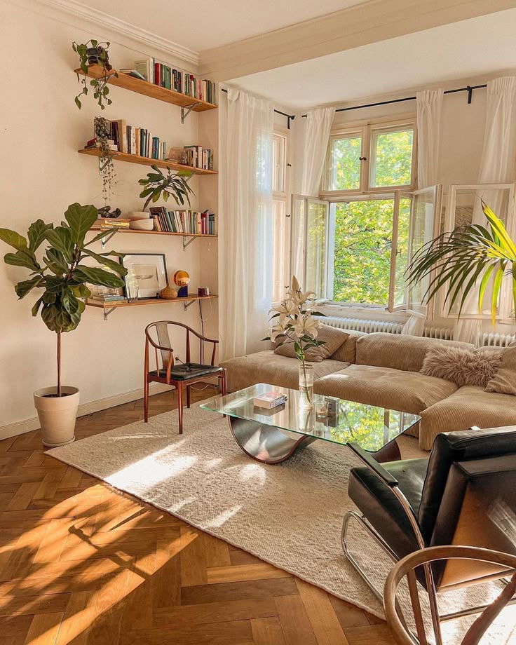 Cozy living room with open windows, lush plants, bookshelves, a glass coffee table, and neutral tones for a serene atmosphere.