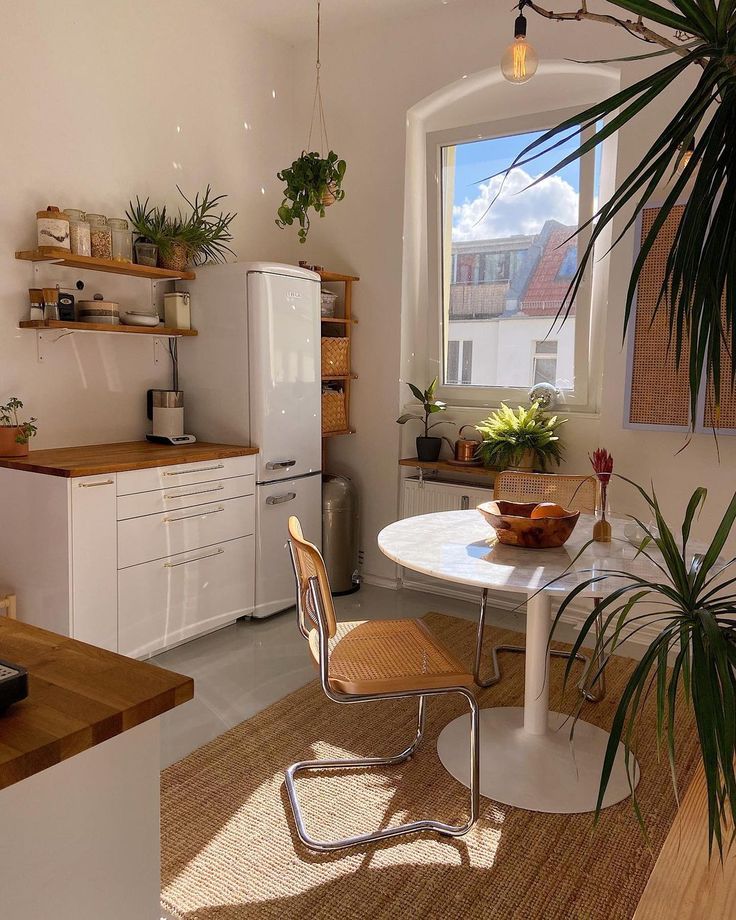 Bright kitchen with wooden counters, hanging plants, white fridge, and round table by sunny window. Cozy, minimalist decor.