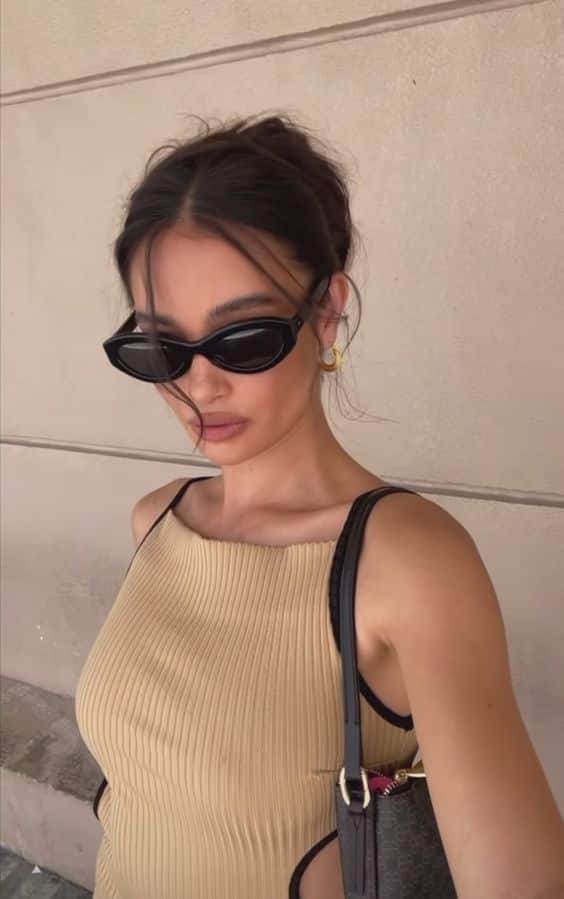 65a76b9f-3df4-47d1-9b8e-5a6538a9d2ea - Handy Home Woman in sunglasses wearing a beige dress poses with a stylish handbag, against a neutral wall background.