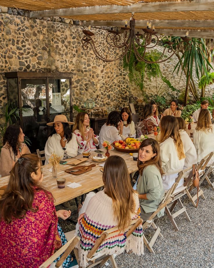 Group of women enjoying a cozy gathering at a rustic outdoor setting with stone walls and plants, seated around a wooden table.