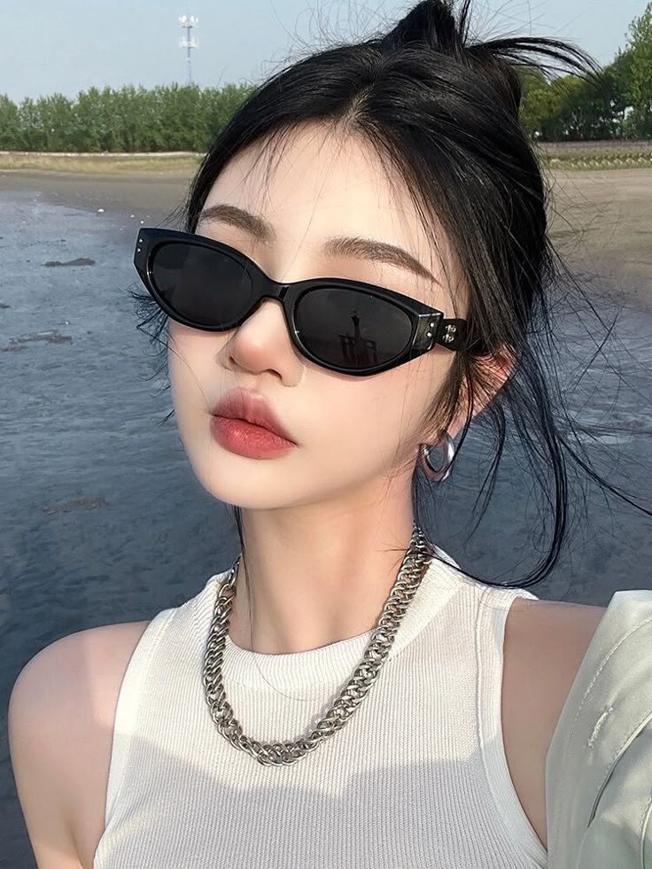 Stylish woman with black sunglasses by the beach, wearing a white top and silver chain necklace.