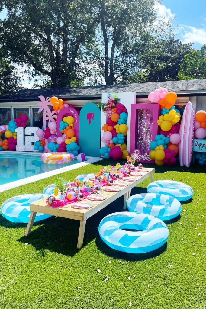 Colorful backyard pool party with vibrant balloon decor, table setup, and inflatable rings on green grass under sunny skies.