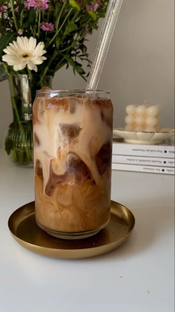 Iced coffee swirled with milk in a glass, next to fresh flowers and books, on a gold tray. Refreshing and stylish beverage.