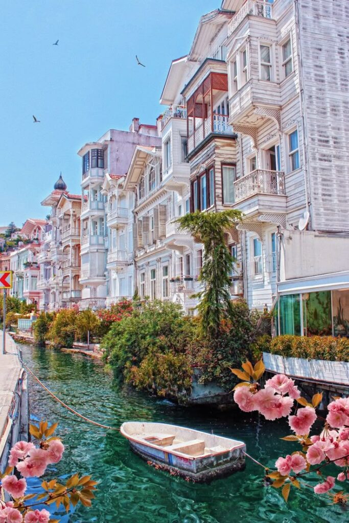 Este fotógrafo está obsesionado con los lugares - Handy Home Charming canal with a rowboat, lined by historic wooden houses and vibrant flowers under a clear blue sky.