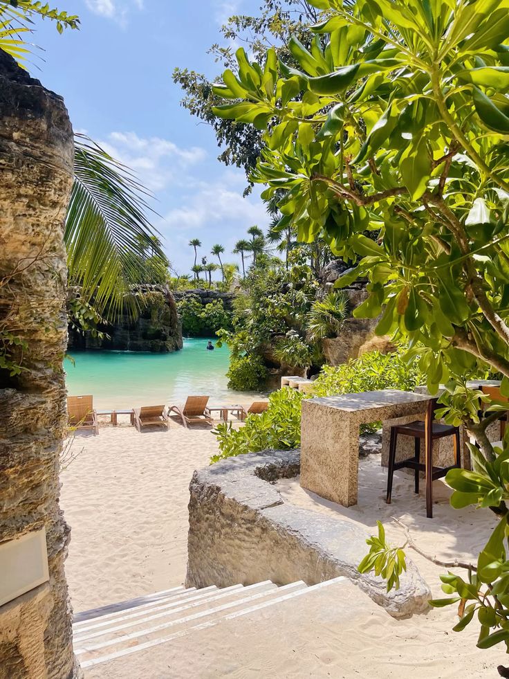 Tropical beach scene with turquoise water, lush greenery, sun loungers, and stone steps leading down to a sandy shore.