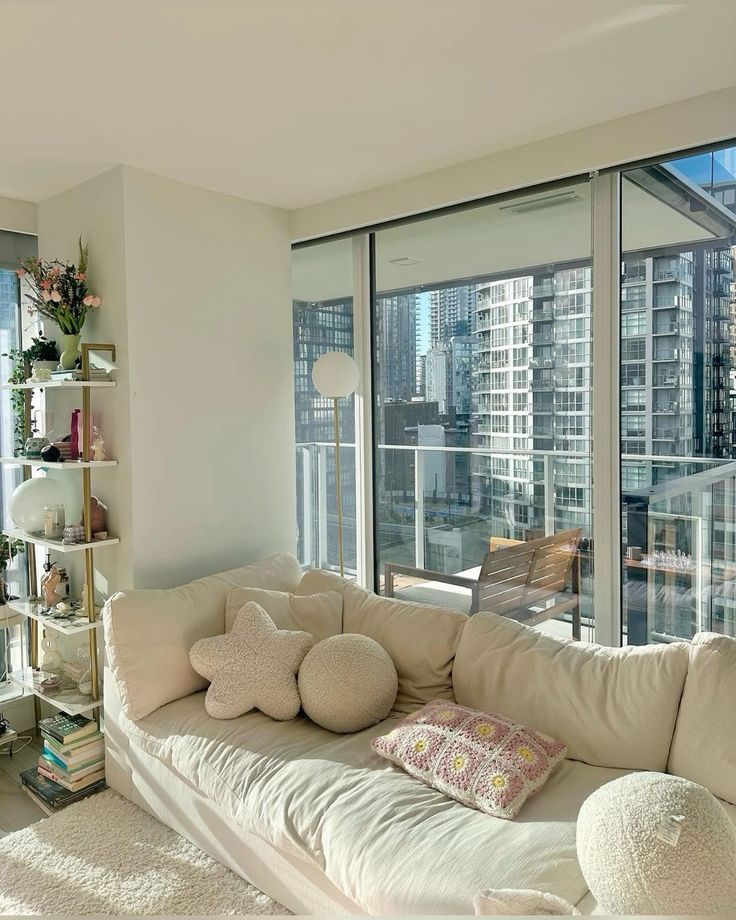 Cozy modern living room with beige sofa, decorative pillows, plants, and large windows showcasing an urban cityscape view. Perfect relaxation spot.