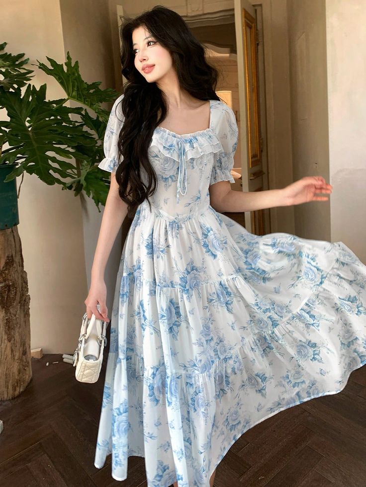 Woman in floral blue and white dress gracefully twirling indoors, holding a small white bag. Elegant fashion and style.