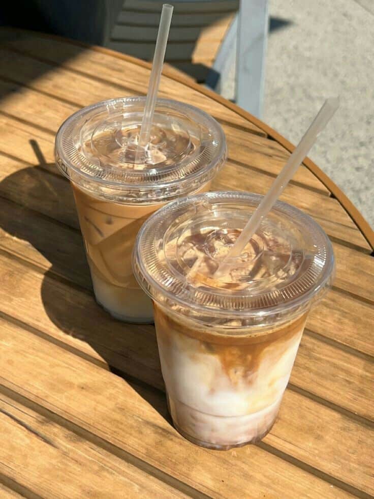 Two iced coffee drinks with clear lids and straws on a sunlit wooden table, perfect for a refreshing summer day.