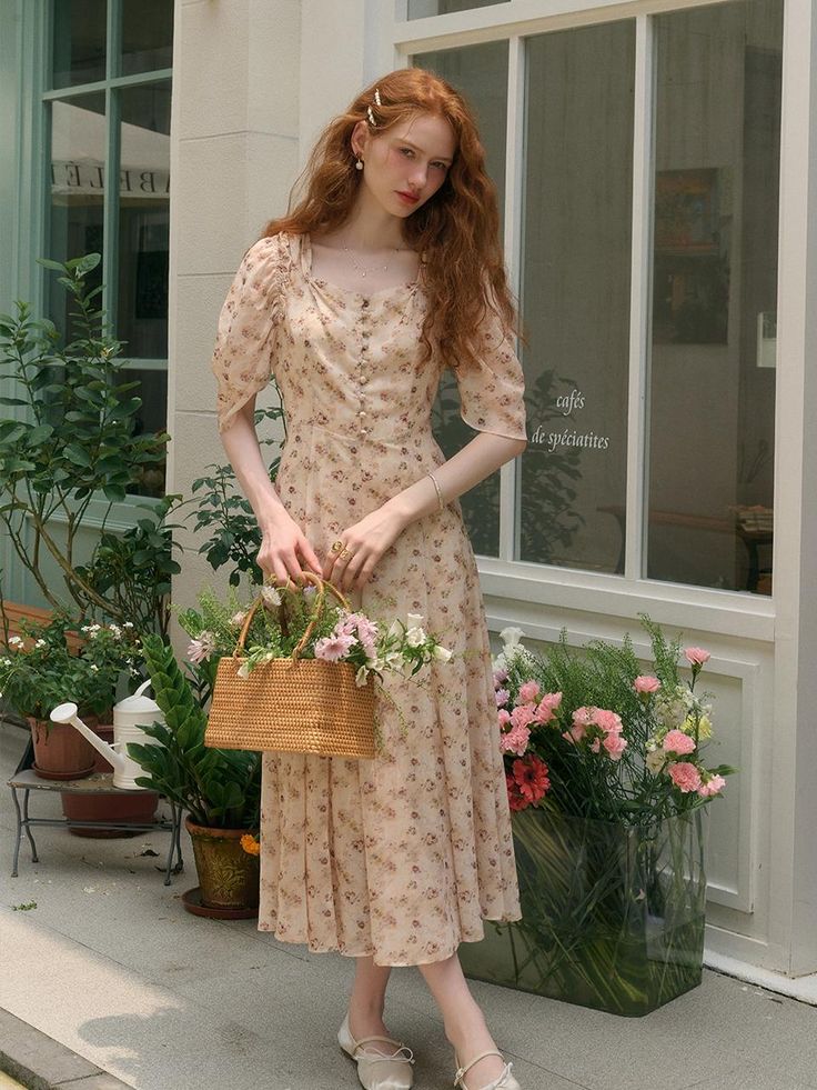 Woman in floral dress holding wicker basket with flowers stands by a cafe window, surrounded by plants.