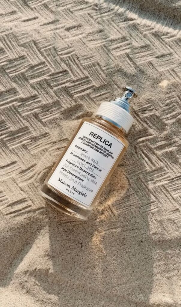 Perfume bottle of Maison Margiela Replica Beach Walk resting on sand, capturing a summer scent with notes of sandalwood and bergamot.