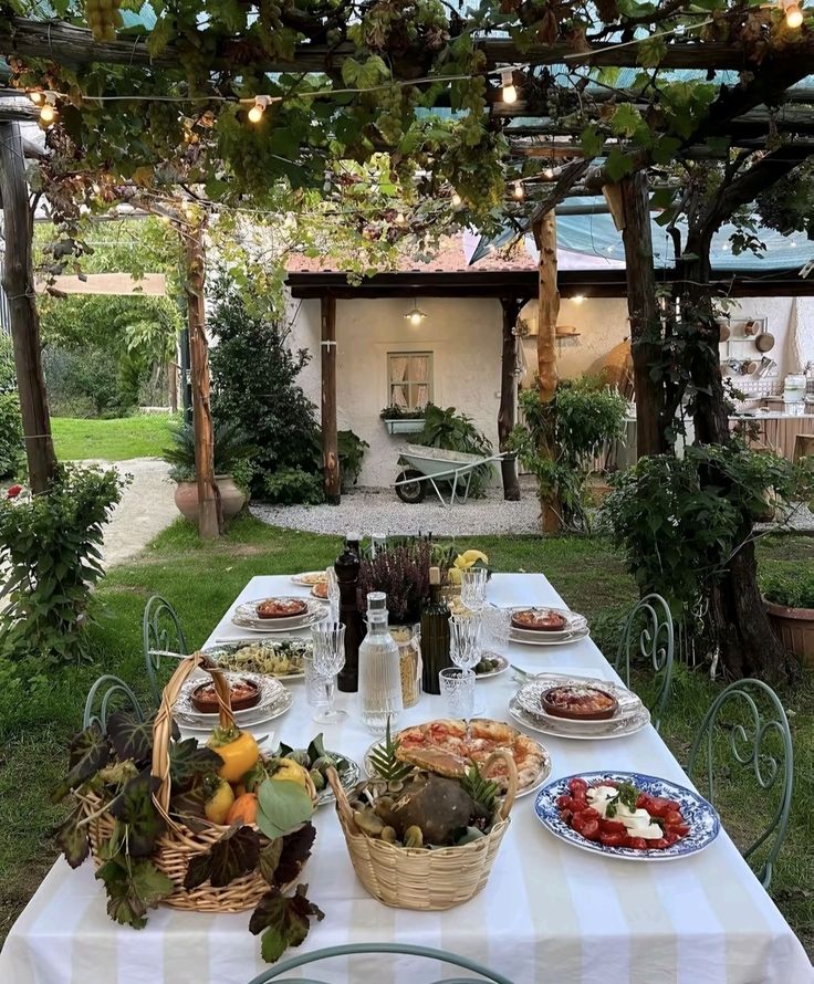 Rustic outdoor dining setting with string lights, featuring a white tablecloth, assorted foods, and lush greenery. Cozy garden ambiance.