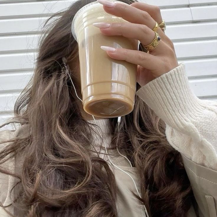 Woman with long hair holds iced coffee while wearing cozy sweater and rings, enjoying a casual day with headphones in.
