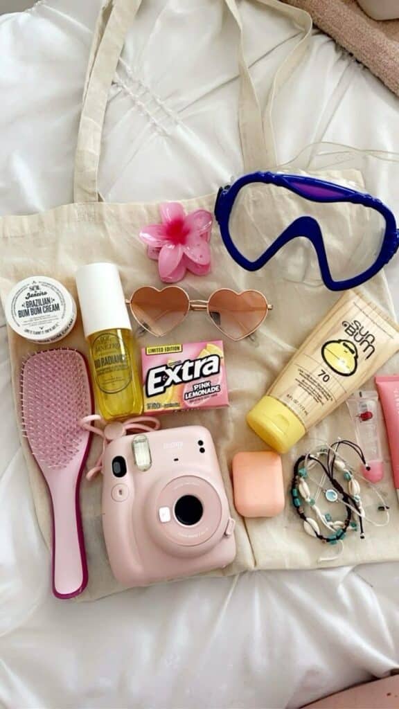 Beach essentials: camera, sunscreen, sunglasses, gum, snorkel, hairbrush, bracelets, and cream on a fabric bag. Fun summer vibes!