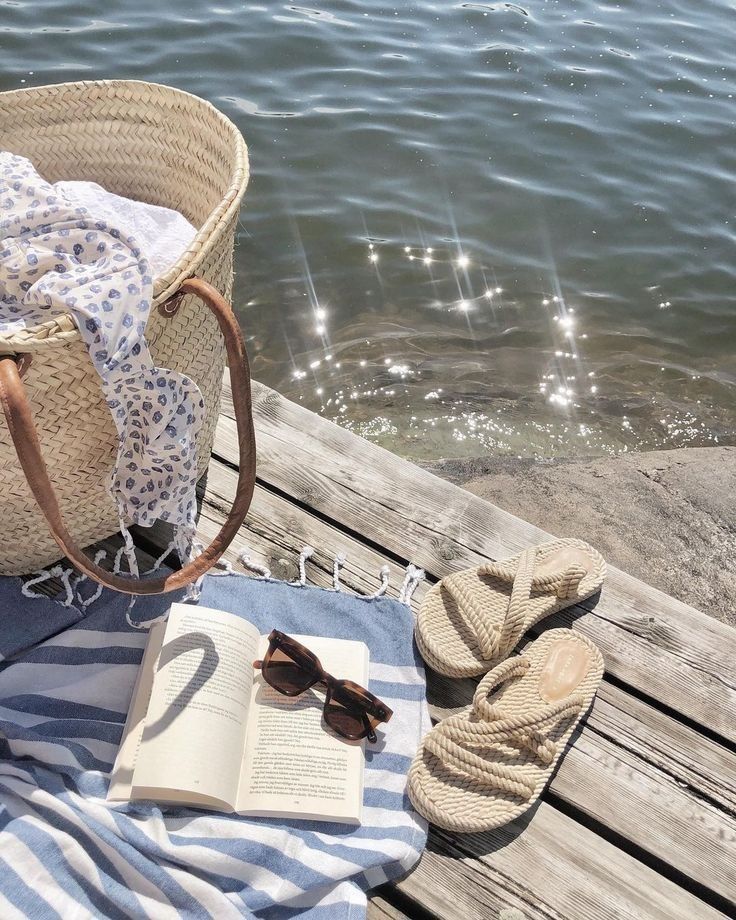 Beach relaxation scene: Woven tote, book, sunglasses, and sandals on a striped towel, next to serene water on a sunny day.