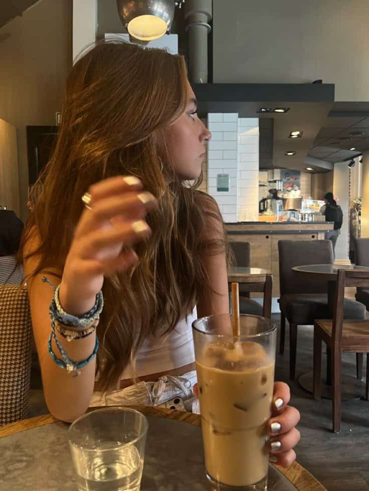 Young woman in a café enjoying an iced coffee, wearing bracelets and surrounded by a cozy modern interior.