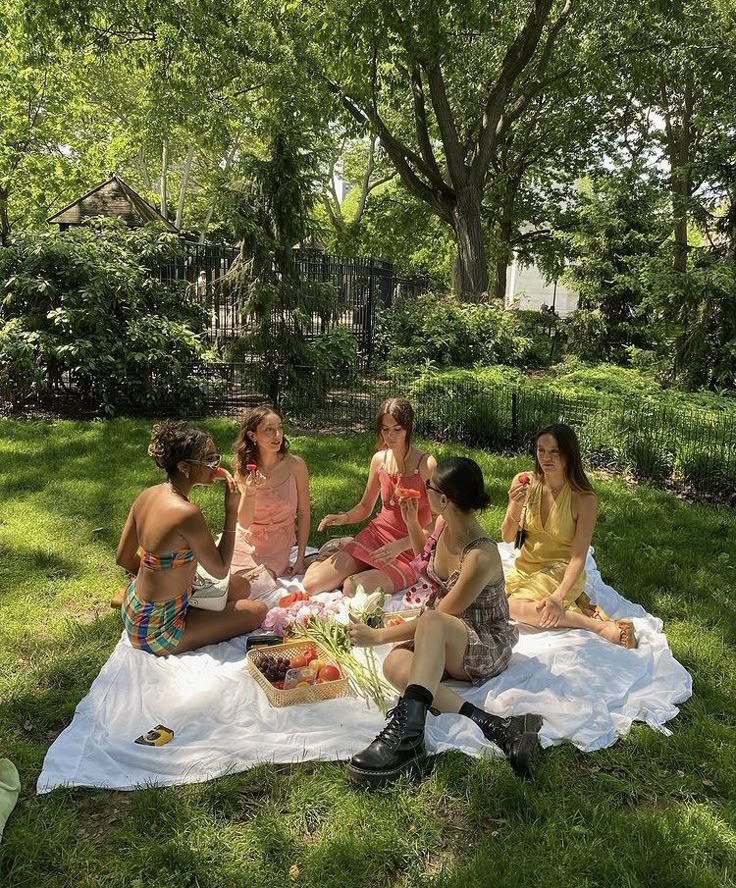 Friends enjoying a summer picnic on a grassy field, surrounded by trees, sharing food and laughs on a sunny day.