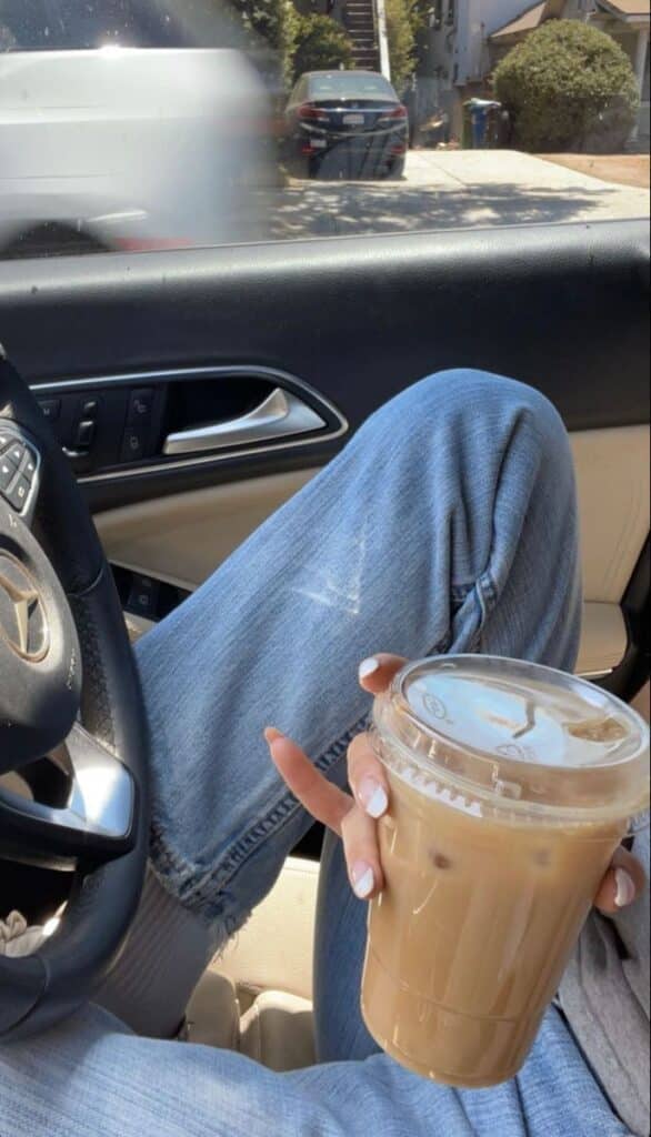 Person in car enjoying iced coffee, wearing jeans, sunny day outside. Steering wheel in view. Relaxed moment.