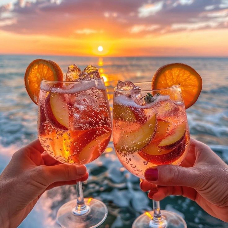 fa43dc25-5460-4002-b77b-a4c44934c0e5 - Handy Home Two vibrant cocktails with fruit slices, raised in a toast at sunset by the ocean, reflecting warm, golden hues on the water.