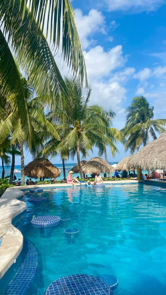 Tropical beach resort with palm trees, vivid blue pool, and straw umbrella huts under a sunny sky. Perfect for a relaxing getaway.