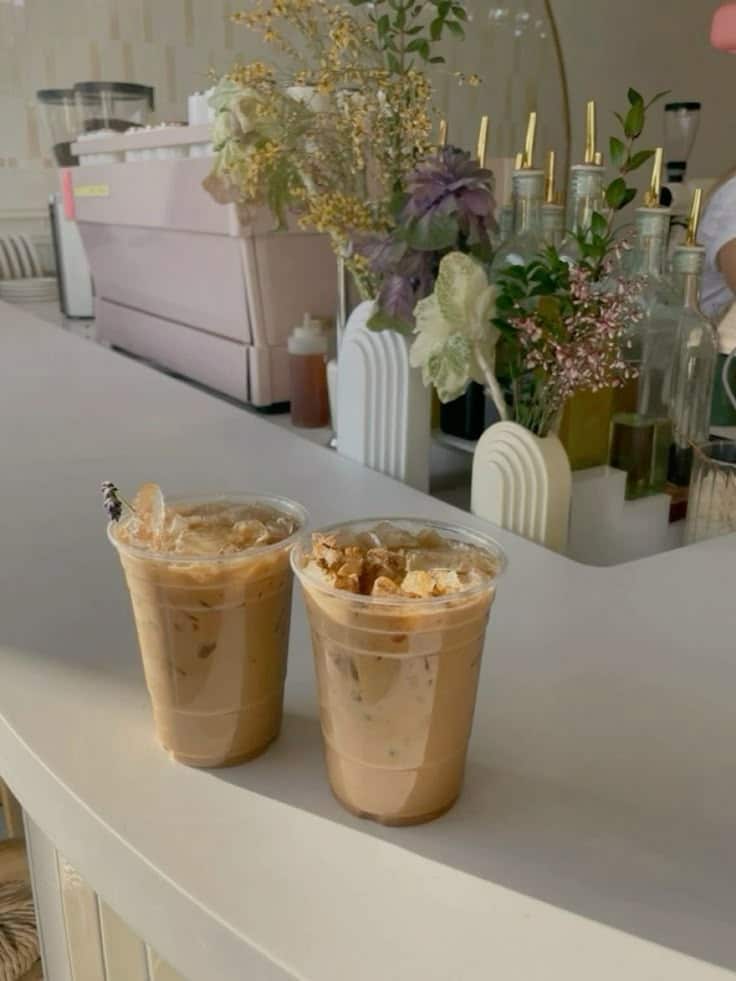 Two iced coffees on a cafe counter with greenery and equipment in the background, creating a cozy and inviting atmosphere.