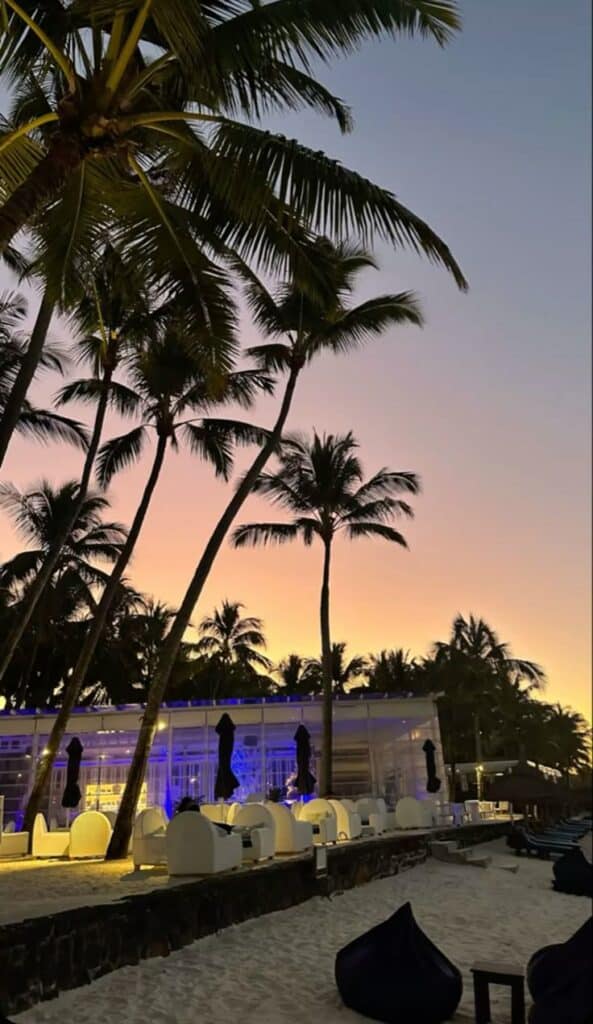 Tropical beach at sunset with palm trees silhouetted against colorful sky, modern seating area and softly lit building in the background.