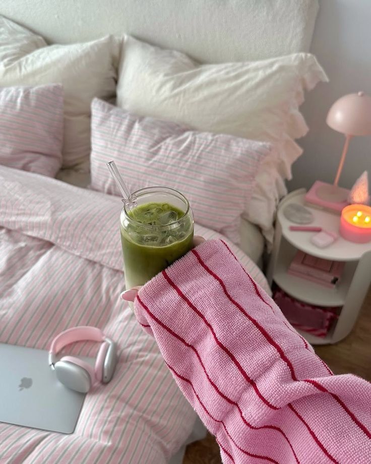 Cozy bedroom scene with pink-striped bedding, iced matcha latte, headphones, and a lit candle on the nightstand.