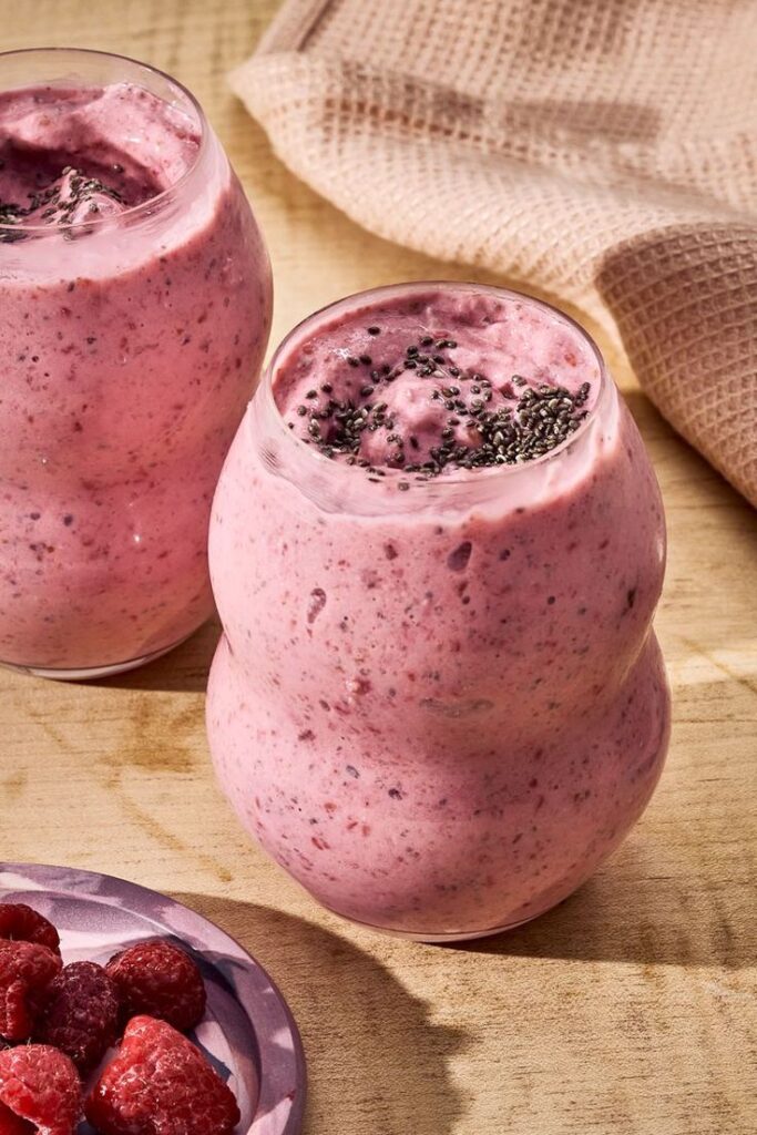 Creamy raspberry smoothie in unique glasses, topped with chia seeds, on a wooden surface with fresh raspberries nearby.