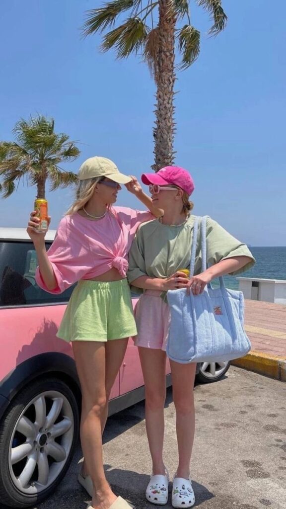 047e1fe7-6cb3-44fb-87d9-e537ddf7dcff - Handy Home Two friends in pastel outfits enjoy a sunny beach day, standing by a pink car with palm trees and ocean in the background.
