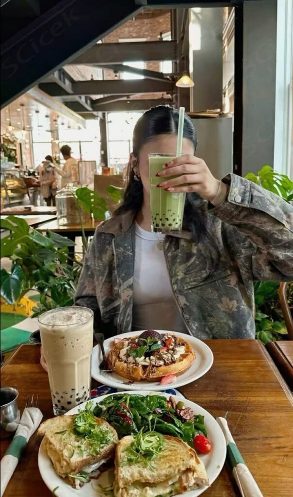 Person holding green bubble tea in cafe, with toast, salad, and another bubble tea on the table. Cozy, delicious brunch setting.