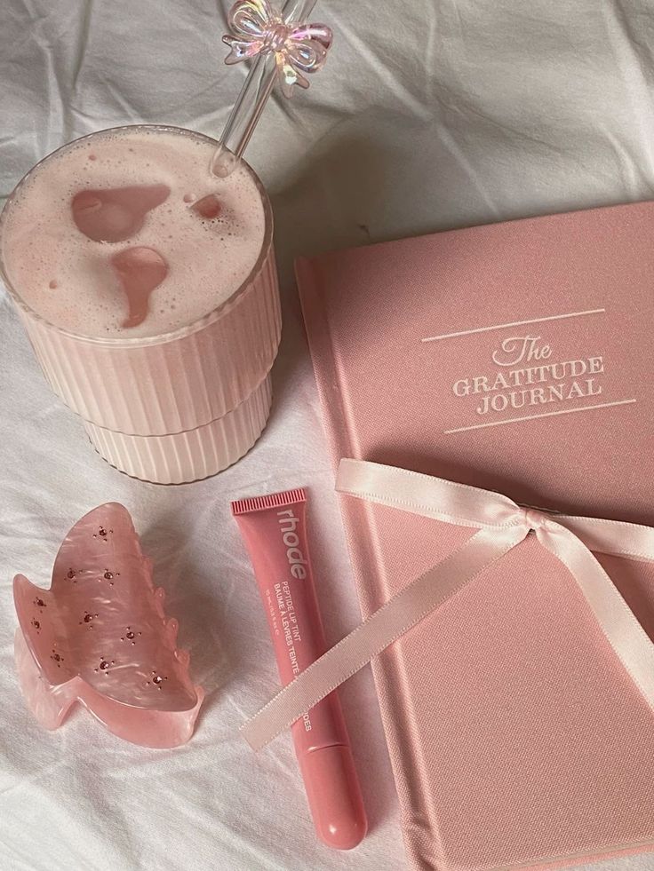 Pink aesthetic flatlay with a gratitude journal, frothy drink, butterfly hair clip, and a pink lip product on a white surface.