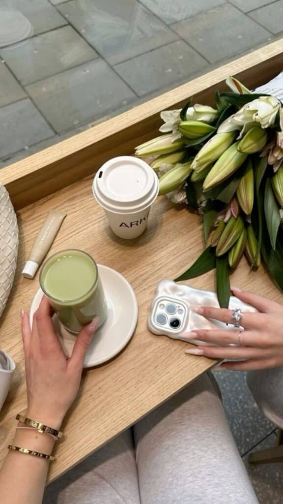 Cozy cafe moment: enjoying a matcha latte with lilies, smartphone, and stylish accessories on a wooden table.