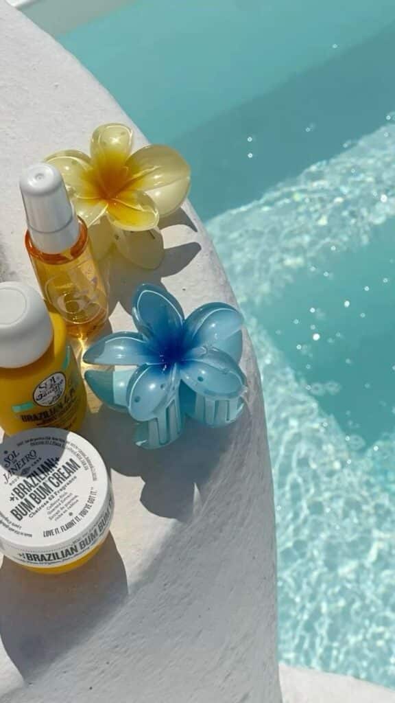 Poolside beauty essentials: tropical-themed hair clips and skincare products on a white ledge by a sparkling blue pool.