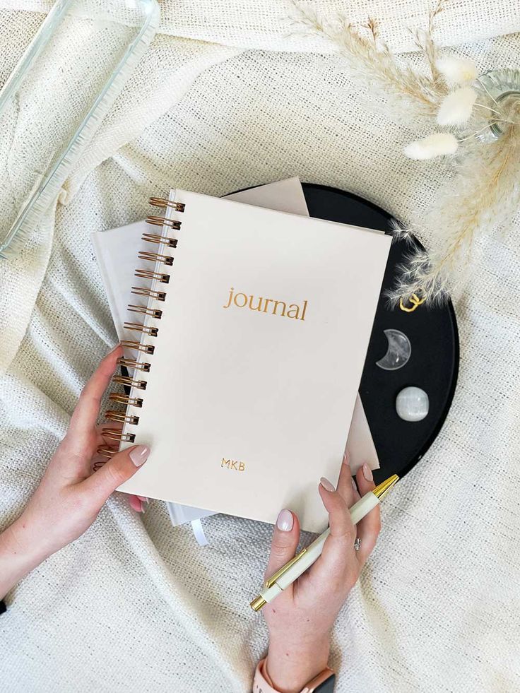 Person holding a spiral journal and pen on a cozy fabric background, with decorative stones and dried flowers nearby.
