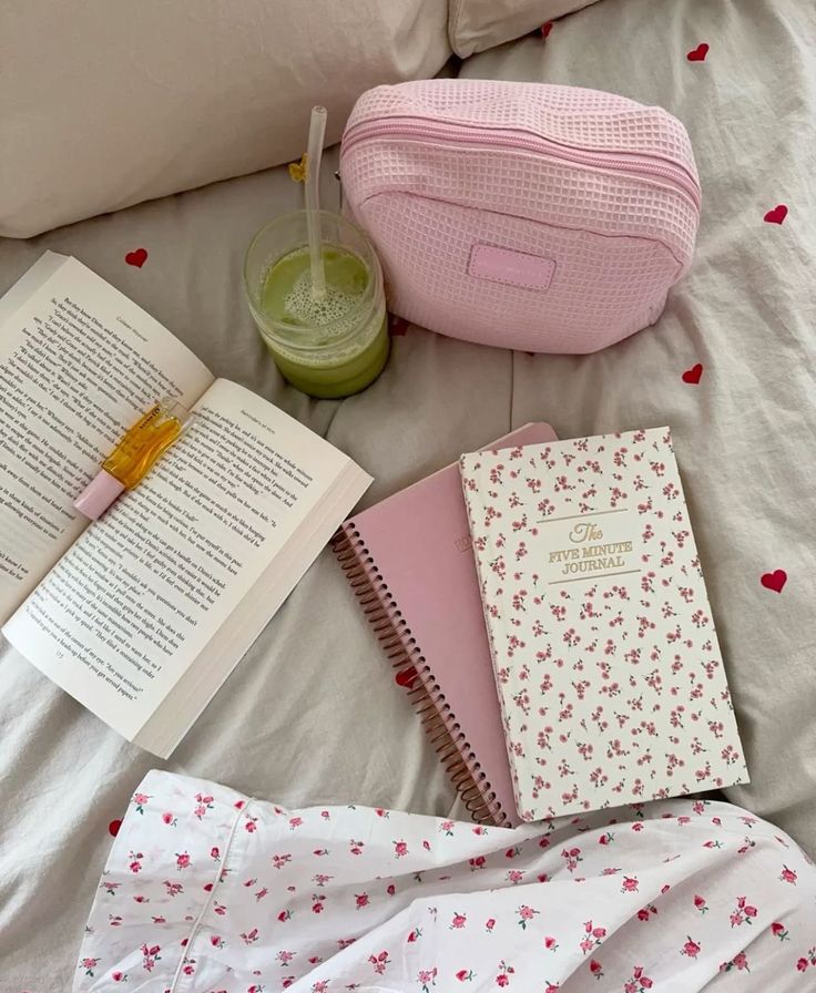 Cozy setup with open book, colorful journal, pink pouch, and green smoothie on a bed with floral sheets, creating a relaxing vibe.