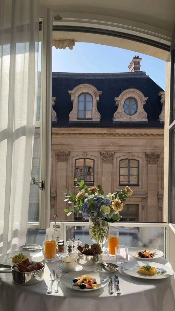 Elegant breakfast setup by a window with a Parisian view, featuring flowers, juice, pastries, and fruits.