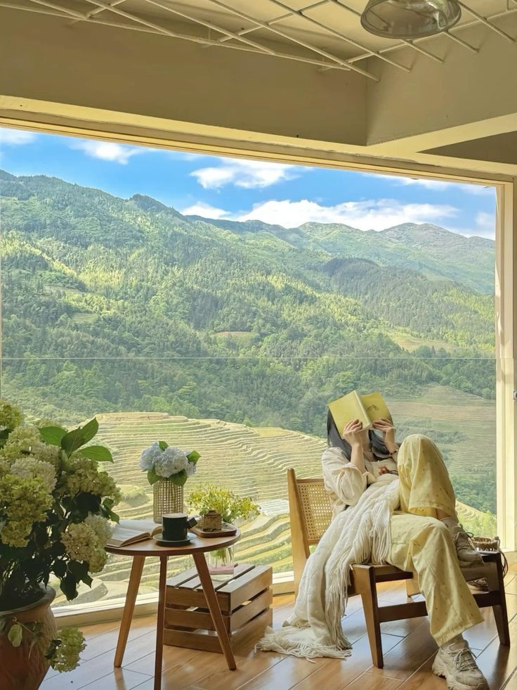 Woman relaxing indoors with a book, overlooking stunning terraced mountains and greenery. Cozy space with hydrangeas and coffee.