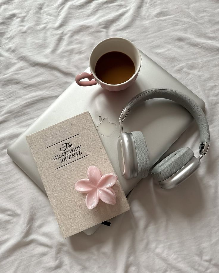 A cup of tea, gratitude journal, headphones, and laptop on a bed. Cozy workspace inspiration for mindfulness and productivity.