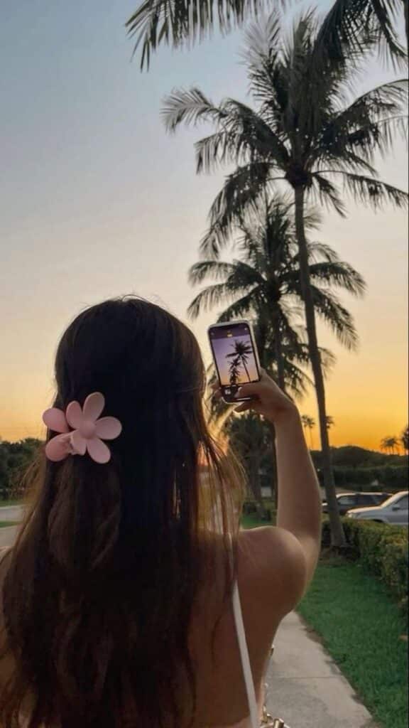 Woman capturing sunset with smartphone, palm trees in view, pink flower hair accessory, vibrant evening sky.